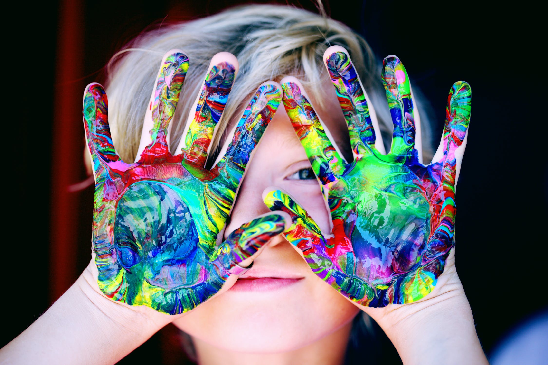 young child covering face with hands dirty from multicolor paint